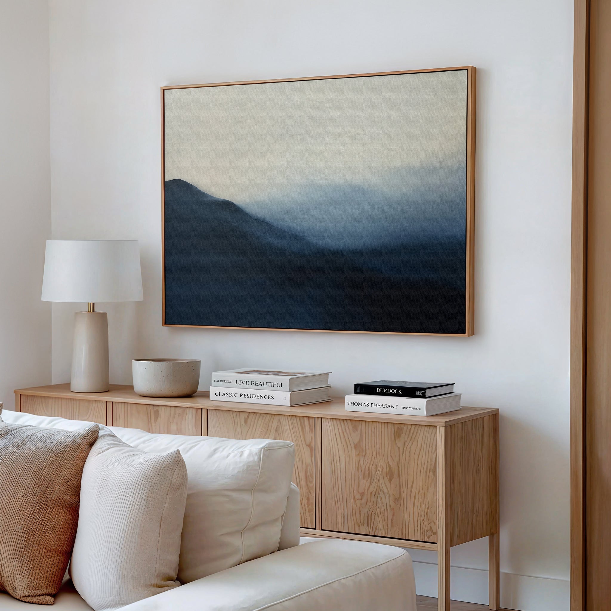 A minimalist living room features a light wooden sideboard, books, a lamp, a bowl, and the Shadowed Valley Canvas Art – Horizontal depicting blue mountains and mist hanging above a white sofa on a white wall.