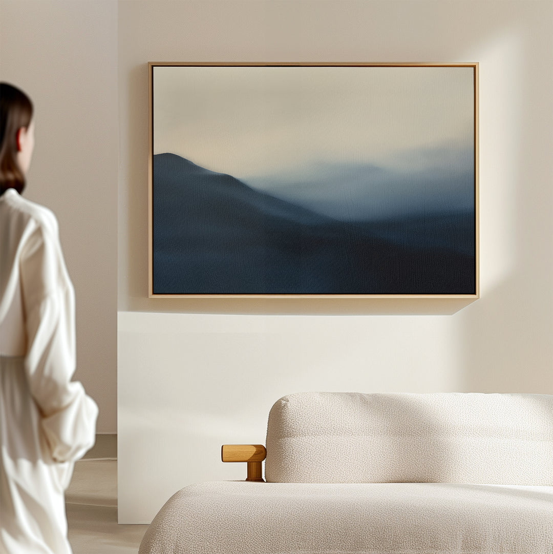 A person in a white shirt stands in a minimal, bright room with calming bedroom decor, featuring a textured beige sofa and the Shadowed Valley Canvas Art – Horizontal, a framed abstract painting of misty blue mountains on the wall.