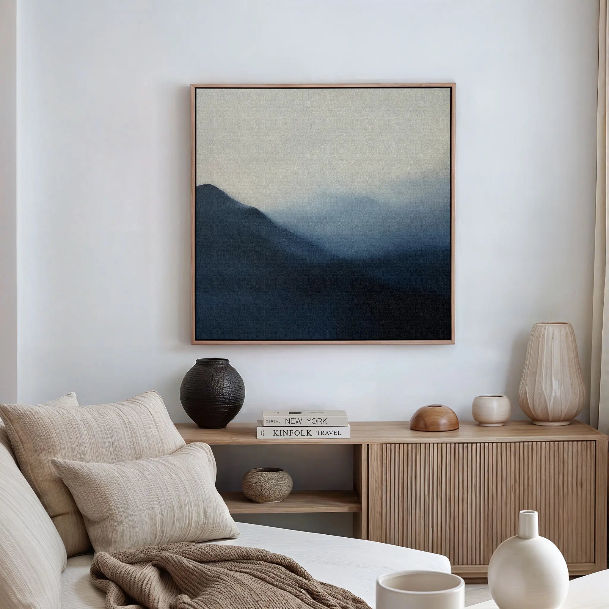 A modern, neutral-toned living room features a beige sofa, light wood sideboard, pottery accents, books, and minimalist decor highlighted by the Shadowed Valley Canvas Art—a framed abstract painting of misty blue mountains on the wall.