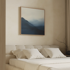 A serene bedroom features a beige upholstered bed with white bedding and pillows. Above, the Shadowed Valley Canvas Art adds minimalist blue mountain vibes. Soft light filters through curtains, with a bare branch on the nightstand.