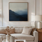 A neutral living room featuring a beige sofa, cream pillows, and calming decor, highlighted by Shadowed Valley Canvas Art—minimalist wall art of blue and gray abstract mountains—plus a side table with white lamp and dried flowers in a vase.