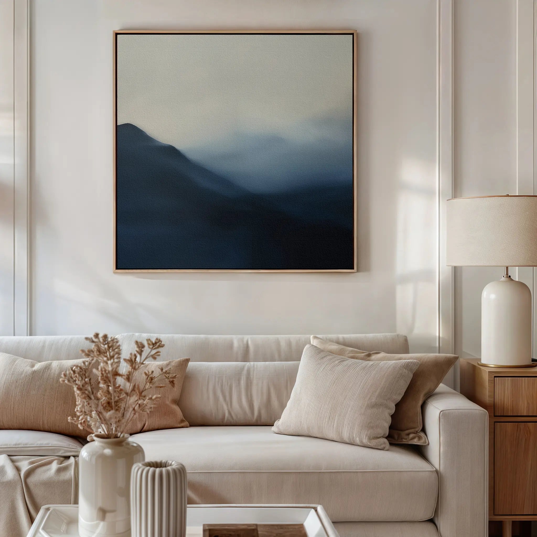 A neutral living room featuring a beige sofa, cream pillows, and calming decor, highlighted by Shadowed Valley Canvas Art—minimalist wall art of blue and gray abstract mountains—plus a side table with white lamp and dried flowers in a vase.