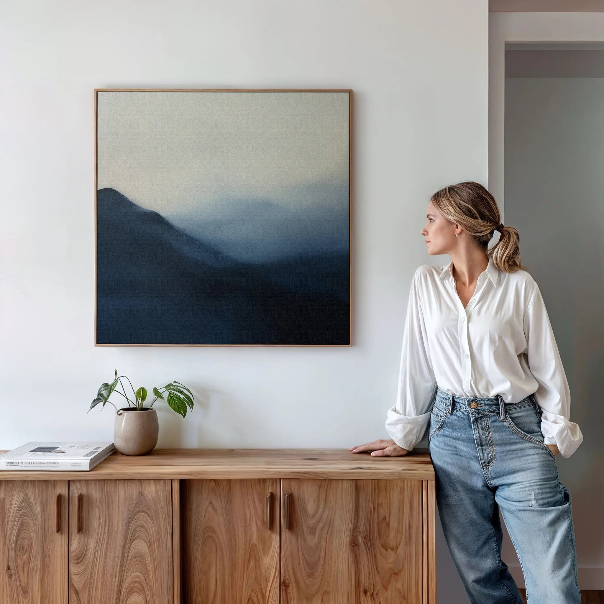 A woman in a white blouse and jeans stands by a wooden cabinet, admiring the Shadowed Valley Canvas Art with blue and gray tones on a white wall. A potted plant and book sit on the cabinet, completing the calming decor of the space.