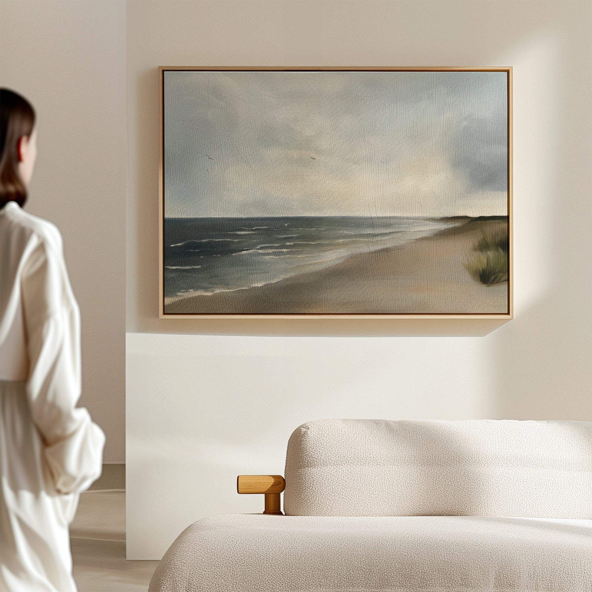 A person in a white shirt stands in a bright, minimal room, admiring Shoreline Calm Canvas Art—a framed beach painting of waves and clouds—displayed above a beige sofa, bringing serene coastal decor to the space.