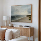 A light wood sideboard with books, a lamp, and a bowl sits against a white wall. Above it hangs the Shoreline Calm Canvas Art, featuring a serene shore and cloudy skies. A white sofa is partially visible in the foreground.