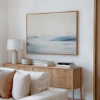A minimal living room with a light wood console table, stacked books, a white lamp, a bowl, and Silent Glaciers Canvas Art—a large wall piece showcasing a tranquil misty blue landscape—displayed above.