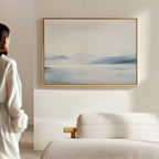 A person stands in a bright, minimal room, admiring Silent Glaciers Canvas Art—minimalist wall art of blue mountains and a misty lake—displayed above a beige sofa.