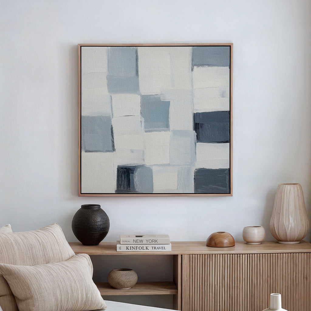 A minimalist living room features the Silent Squares Abstract Canvas Art—blue, gray, and white rectangles above a wooden sideboard—paired with vases, books, and a cushion on a beige sofa for sleek wall art décor.