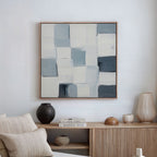A minimalist living room features the Silent Squares Abstract Canvas Art—blue, gray, and white rectangles above a wooden sideboard—paired with vases, books, and a cushion on a beige sofa for sleek wall art décor.