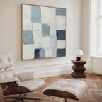 A modern living space with a cream lounge chair and ottoman, a geometric wooden side table and white lamp, herringbone wood floors, and the Silent Squares Abstract Canvas Art in blue, gray, and cream tones displayed above.