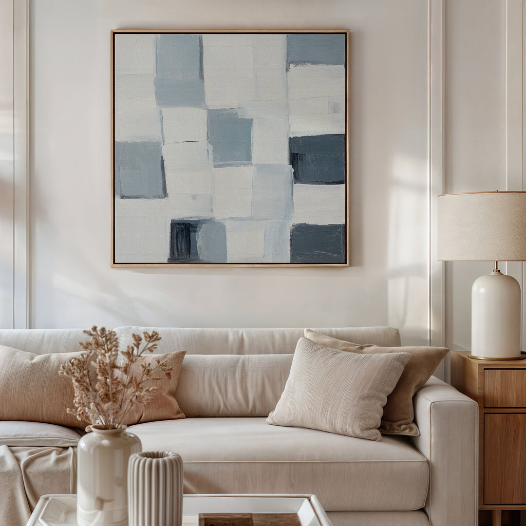 A modern living room features a beige sofa with light cushions, the Silent Squares Abstract Canvas Art above. On the coffee table are a ceramic vase with dried flowers and a ribbed vase; a lamp sits on the wooden side table.