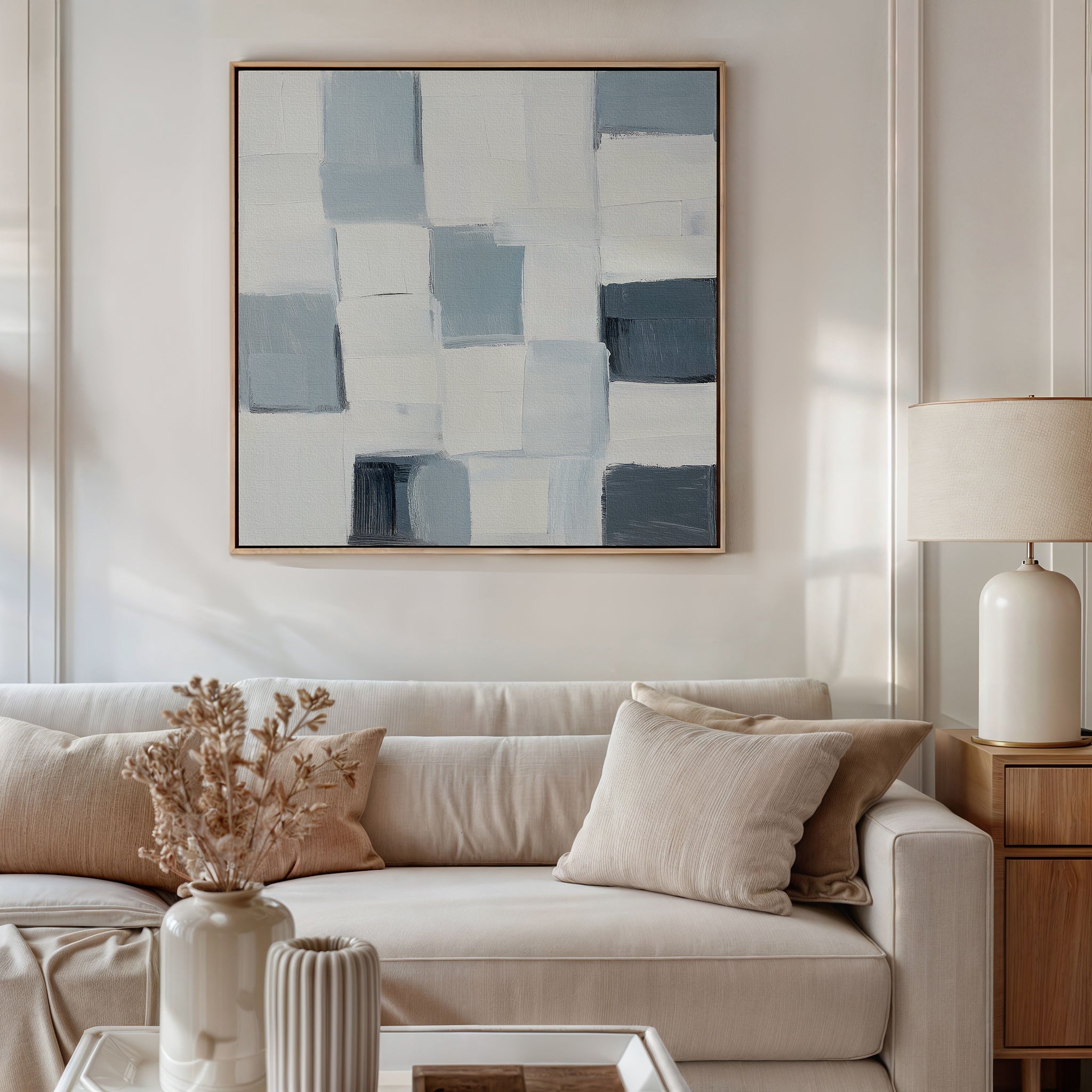 A modern living room features a beige sofa with light cushions, the Silent Squares Abstract Canvas Art above. On the coffee table are a ceramic vase with dried flowers and a ribbed vase; a lamp sits on the wooden side table.