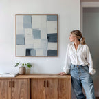 A woman in a white blouse and jeans leans against a wooden cabinet, admiring the Silent Squares Abstract Canvas Art featuring blue, gray, and white blocks. A small plant and a book rest beneath this modern decor accent.