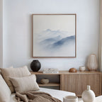 A minimalist living room features a neutral sofa, beige and white cushions, a wooden sideboard with vases and books, and Silent Summits Canvas Art—a framed blue misty mountain scene—bringing serene elegance to your decor.