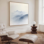 A modern, minimalist living room features a white leather lounge chair with ottoman, wooden floors, a geometric side table with lamp, and Silent Summits Canvas Art—a large painting of misty blue mountains—on the wall.