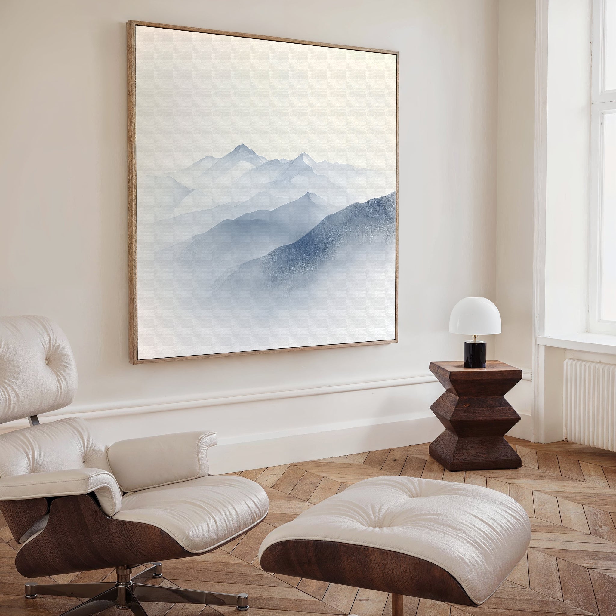 A modern, minimalist living room features a white leather lounge chair with ottoman, wooden floors, a geometric side table with lamp, and Silent Summits Canvas Art—a large painting of misty blue mountains—on the wall.