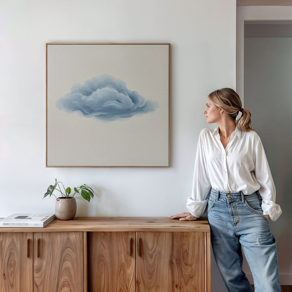 A woman in a white blouse and jeans admires the Single Cloud Canvas Art above a wooden cabinet, which holds a potted plant and book, enhancing the modern decor of the minimalist room.