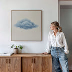 A woman in a white blouse and jeans admires the Single Cloud Canvas Art above a wooden cabinet, which holds a potted plant and book, enhancing the modern decor of the minimalist room.
