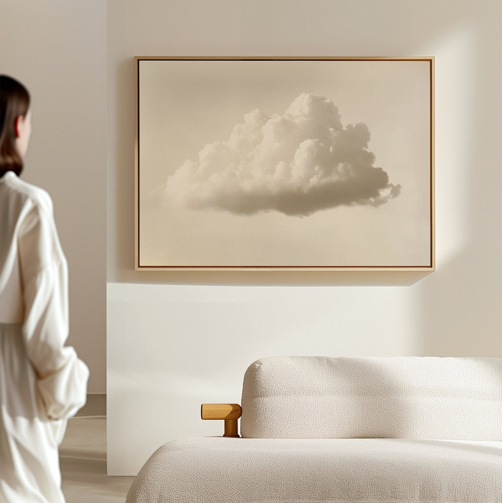 A woman stands in a bright, minimal room, gazing at the Singular Vapor Canvas Art – Horizontal—a framed print of a single cloud—displayed above a beige sofa, evoking calm and simplicity.