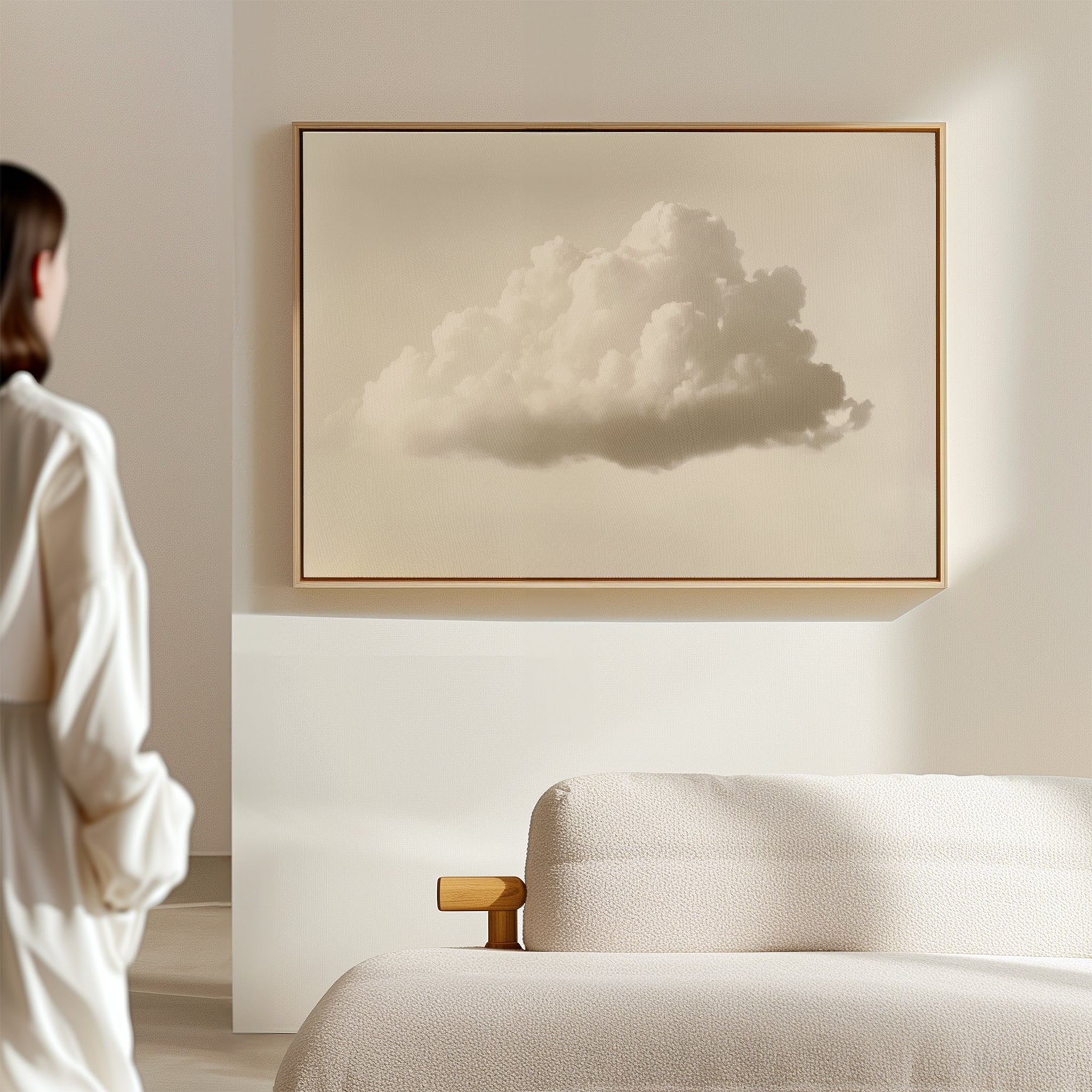 A woman stands in a bright, minimal room, gazing at the Singular Vapor Canvas Art – Horizontal—a framed print of a single cloud—displayed above a beige sofa, evoking calm and simplicity.
