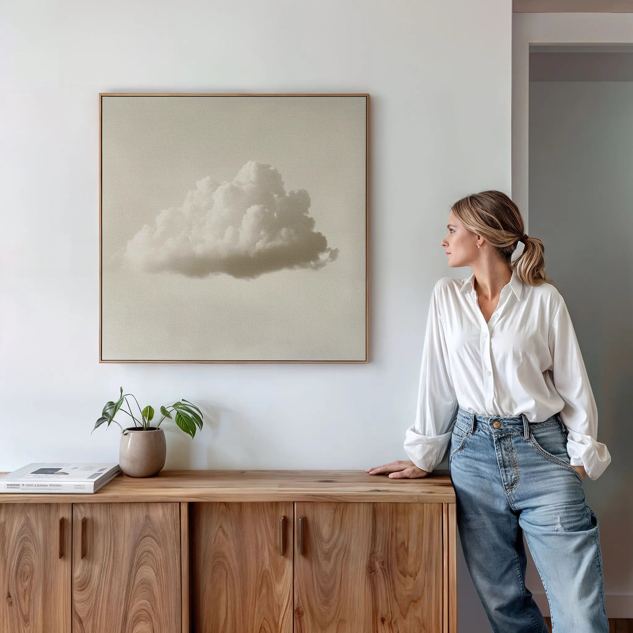 A woman in a white blouse and jeans leans against a wooden cabinet, gazing at the Singular Vapor Canvas Art above her. A small plant and books add subtle style, creating a calm, modern scene ideal for contemporary interiors.