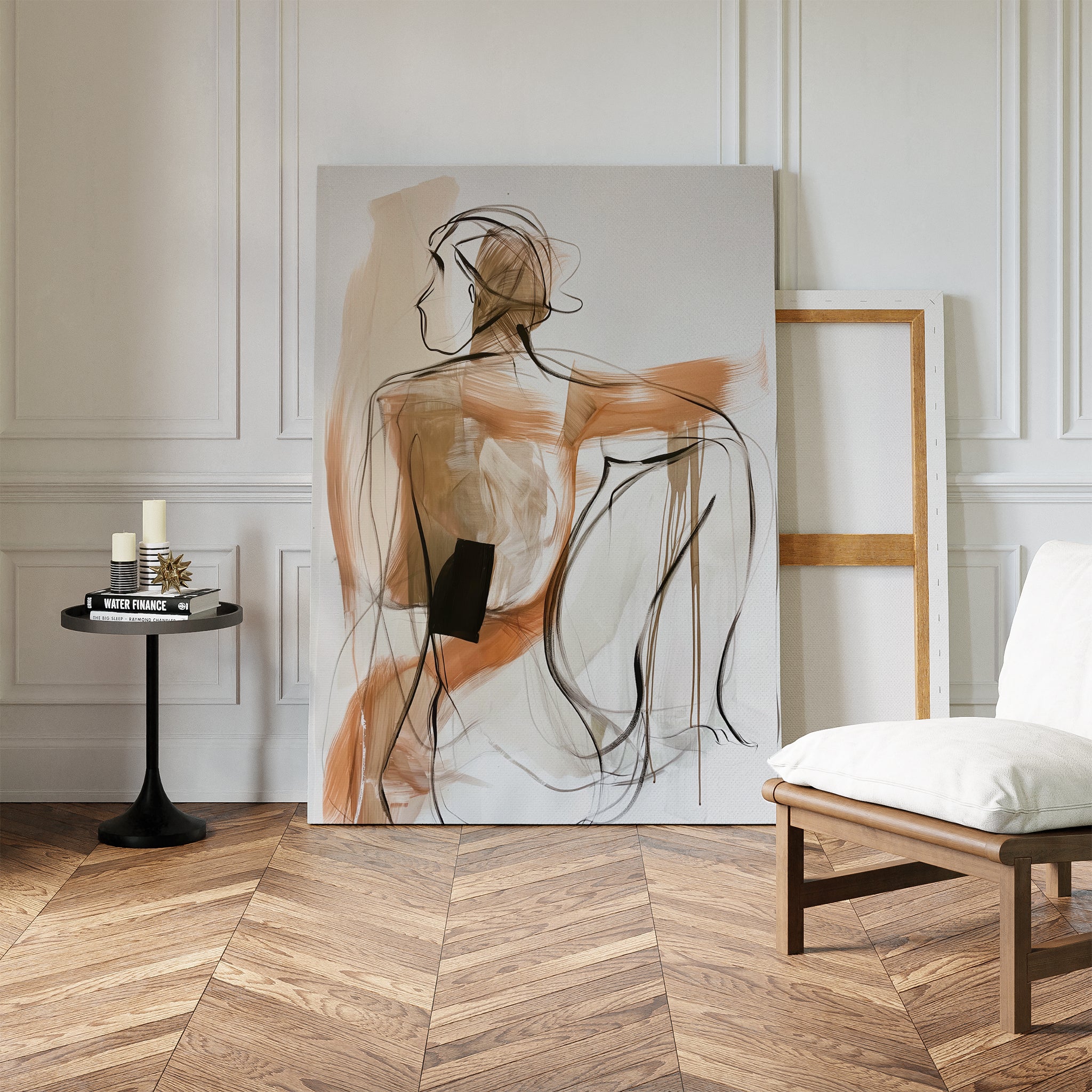 The Sitting Silhouette Abstract Canvas Art leans against a white paneled wall in a stylish room with herringbone wood floors, a low chair, and a black side table decorated with candles, books, and contemporary décor.