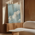 The Sky Serenade Abstract Canvas Art, featuring fluffy white clouds against a blue sky, is displayed on a wooden sideboard in a sunlit room with light wood walls, a textured white sofa, and a round white table.