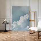 Sky Serenade Abstract Canvas Art, with white clouds in a blue sky, leans against the wall of a minimalist room featuring herringbone wood floors, a modern chair, candles on a table, and an empty wooden frame—ideal for living room decor.