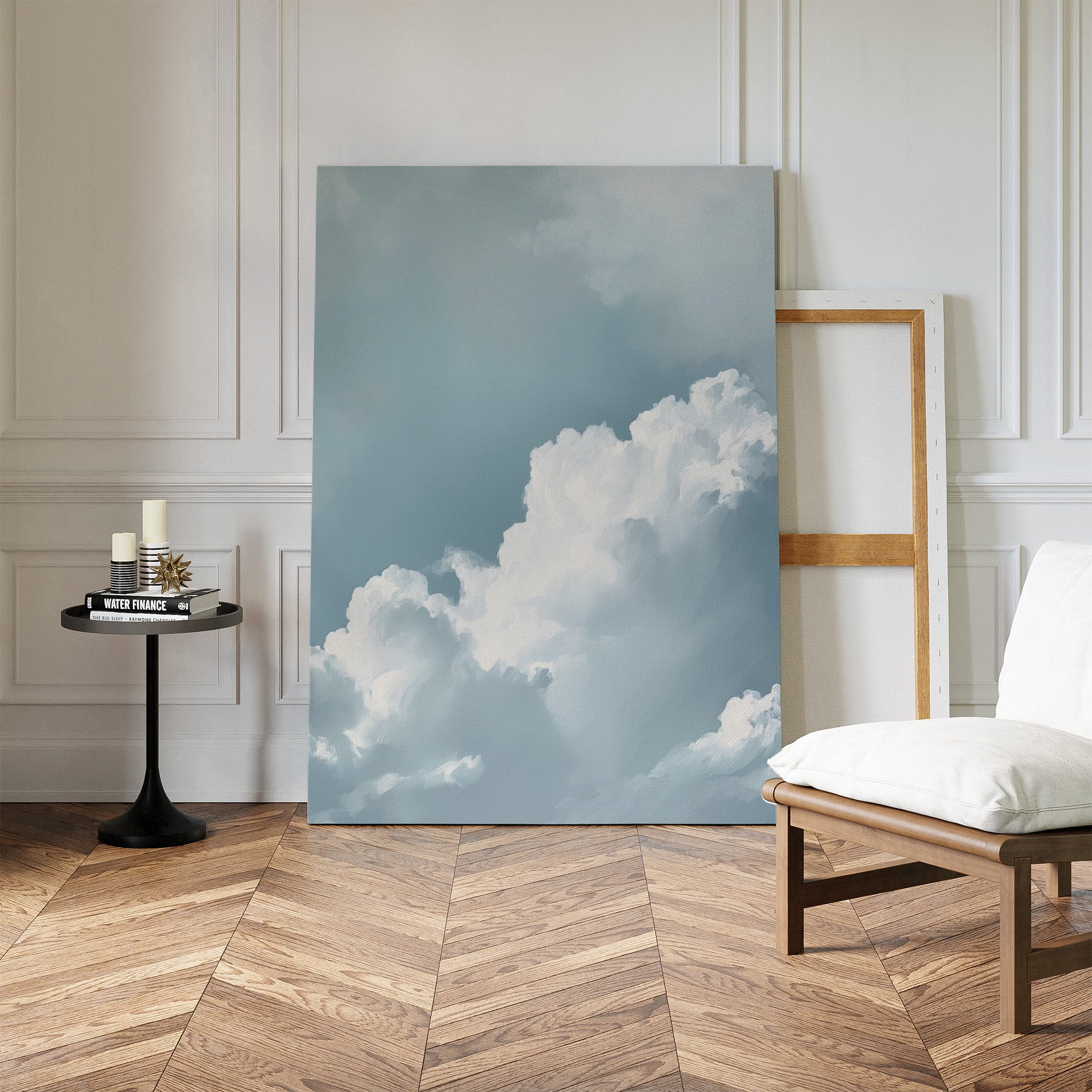 Sky Serenade Abstract Canvas Art, with white clouds in a blue sky, leans against the wall of a minimalist room featuring herringbone wood floors, a modern chair, candles on a table, and an empty wooden frame—ideal for living room decor.