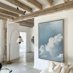 A modern living room with exposed wooden beams, white walls, and Sky Serenade Abstract Canvas Art depicting a blue sky and clouds. A curved floor lamp and white sofa complete the decor, enhanced by natural light from the next room.