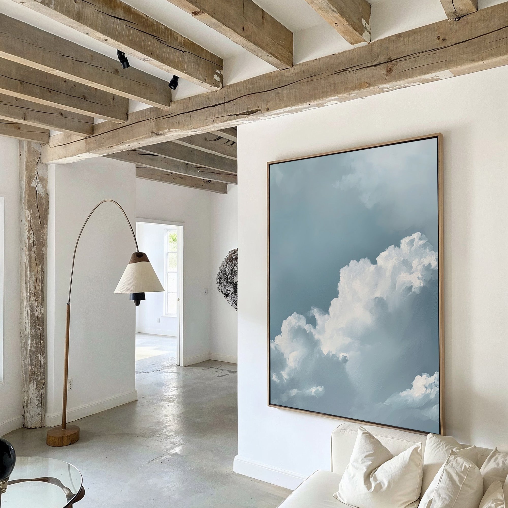 A modern living room with exposed wooden beams, white walls, and Sky Serenade Abstract Canvas Art depicting a blue sky and clouds. A curved floor lamp and white sofa complete the decor, enhanced by natural light from the next room.