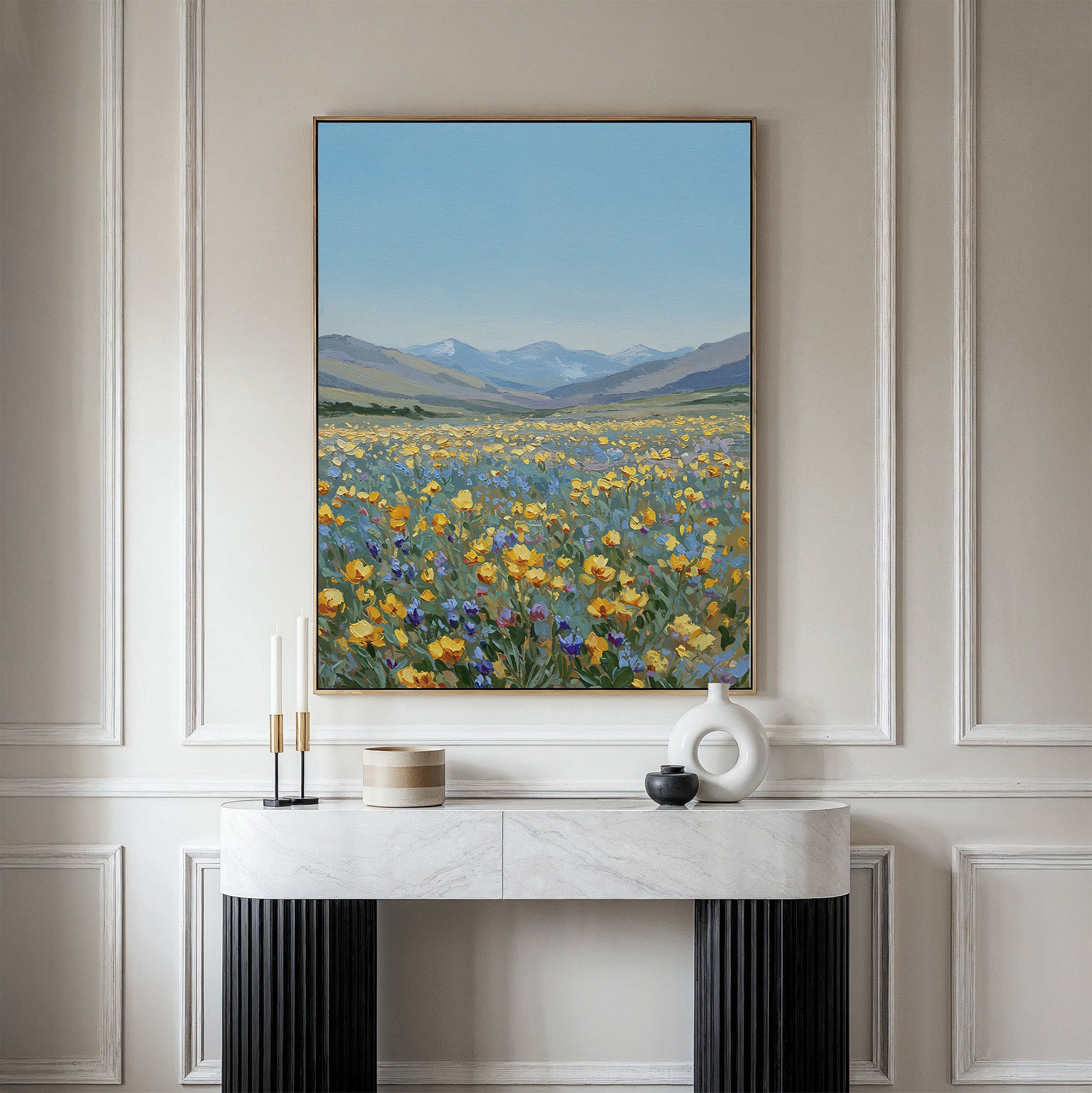 The Skyview Field Canvas Art, featuring a vibrant wildflower field with distant mountains and blue sky, hangs above a modern white and black console table in an elegant paneled room with minimalist decor.