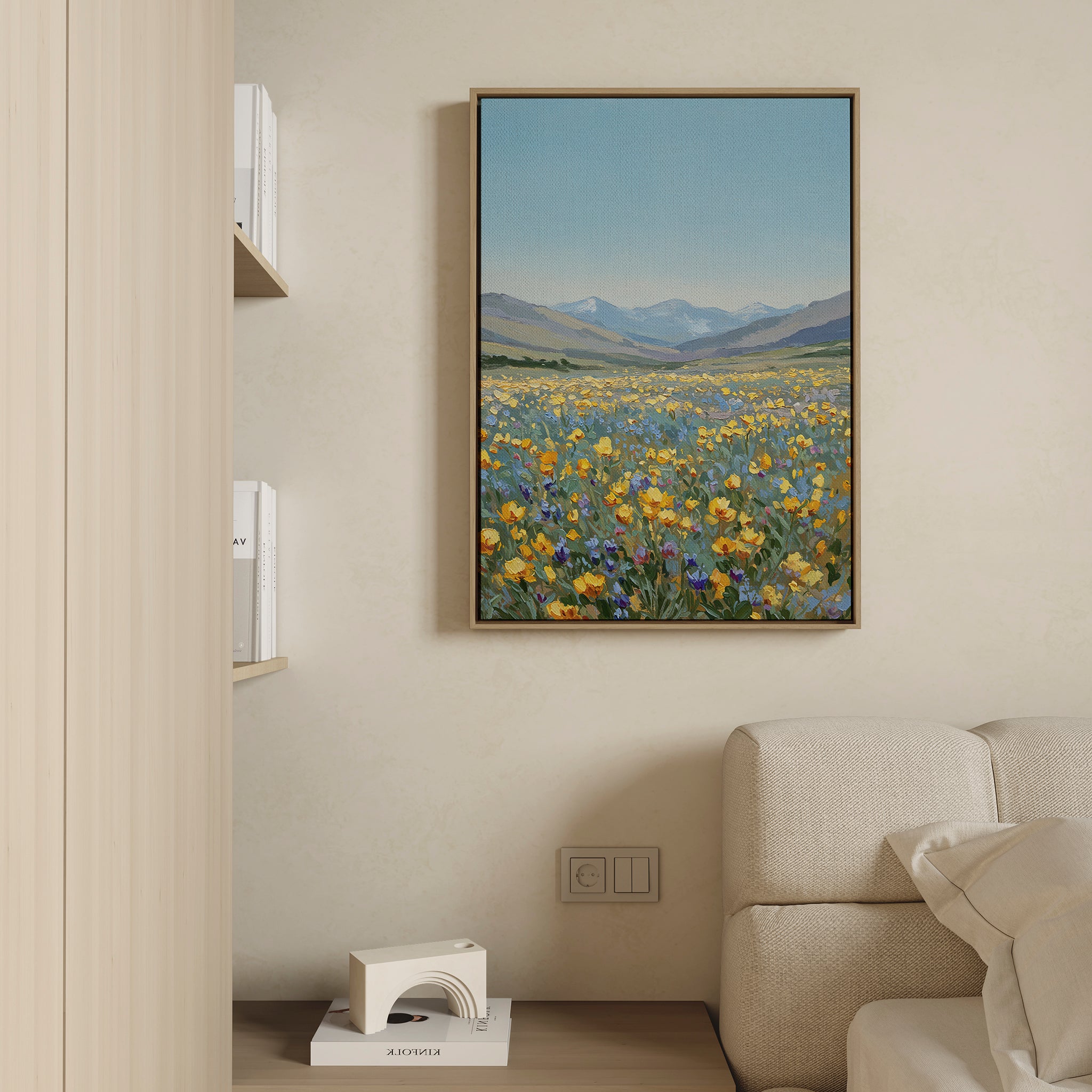 The Skyview Field Canvas Art, featuring a scenic field with yellow and purple wildflowers and distant mountains, hangs above a light sofa for a serene touch—perfect living room or bedroom decor. A shelf beside it displays books and a decorative object.
