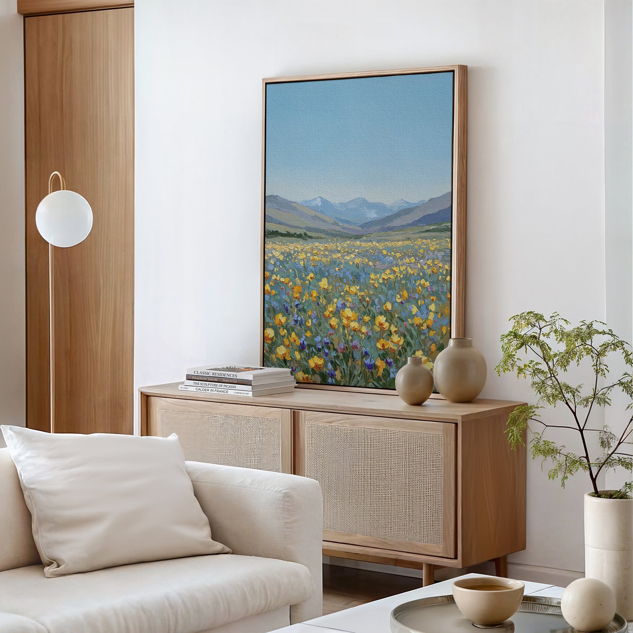 A modern living room with a light wood sideboard, neutral decor, a white sofa, a green potted plant, and large Skyview Field Canvas Art featuring a wildflower field on the wall.