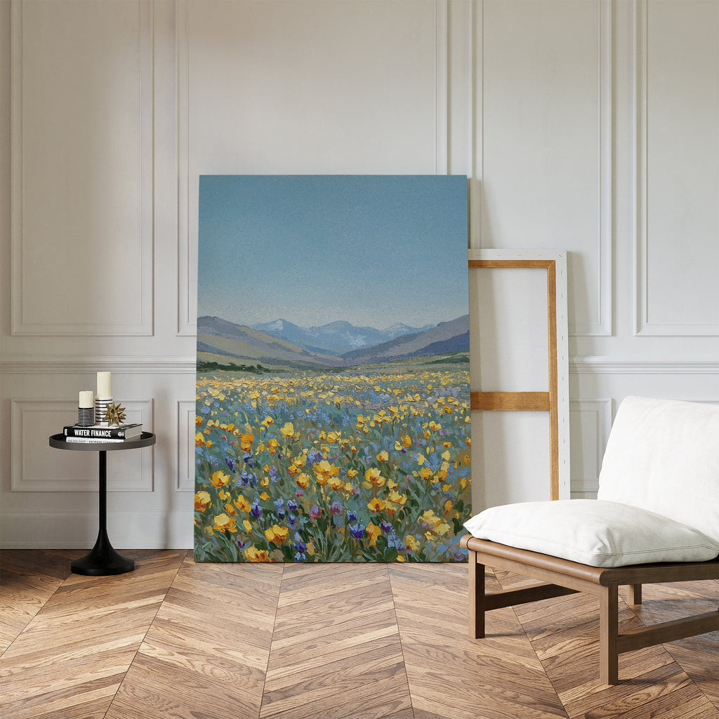The Skyview Field Canvas Art, depicting a wildflower field and mountains, rests against a white-paneled wall, bringing serene minimalist style to cozy bedroom decor, beside a cushioned chair, candlelit side table, and empty frame.
