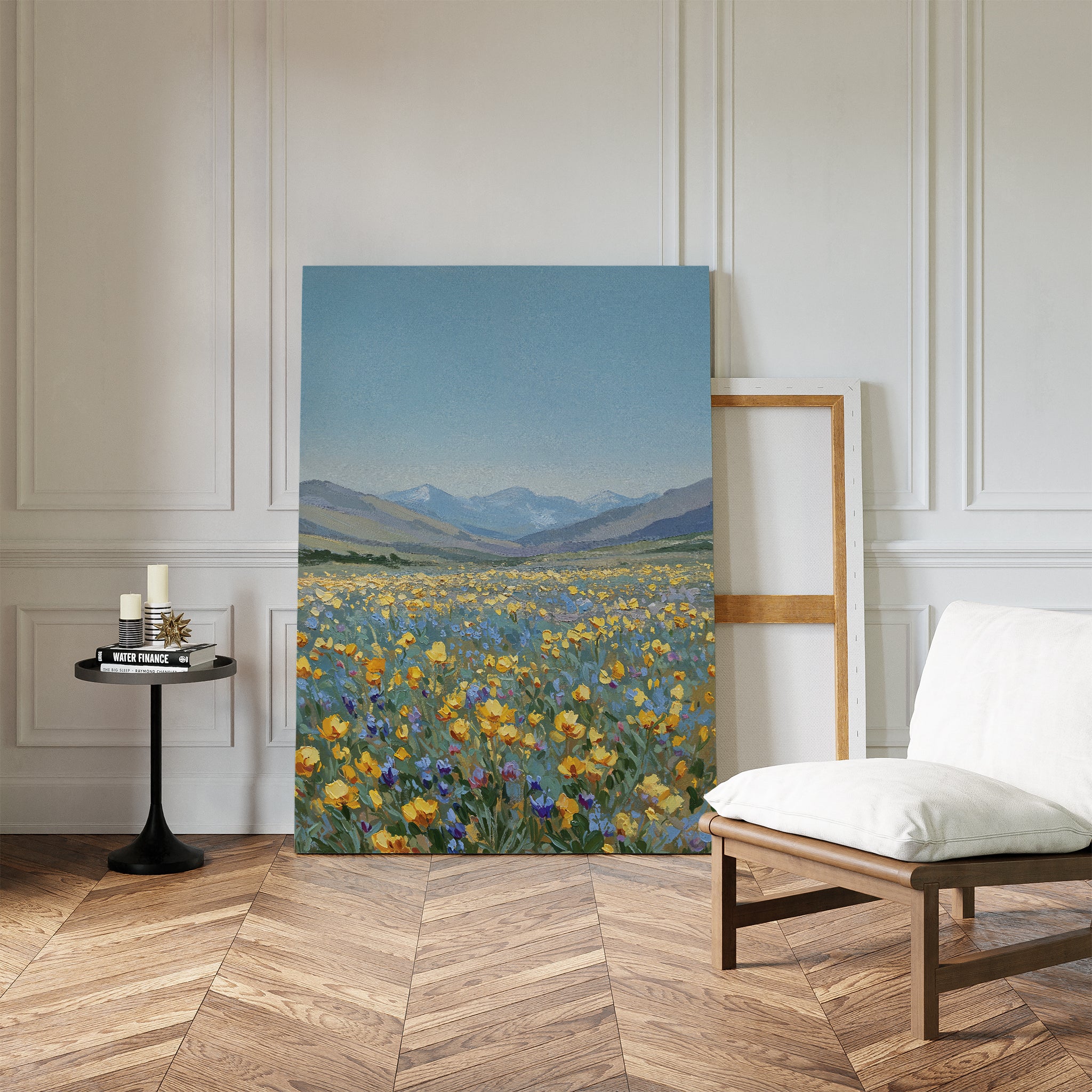 The Skyview Field Canvas Art, depicting a wildflower field and mountains, rests against a white-paneled wall, bringing serene minimalist style to cozy bedroom decor, beside a cushioned chair, candlelit side table, and empty frame.