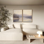 A minimalist living room features a light beige sectional, a small wooden coffee table with a lamp and book, a potted tree, and the Smoky Peaks Set of 2 landscape wall art canvases depicting mountains and water.
