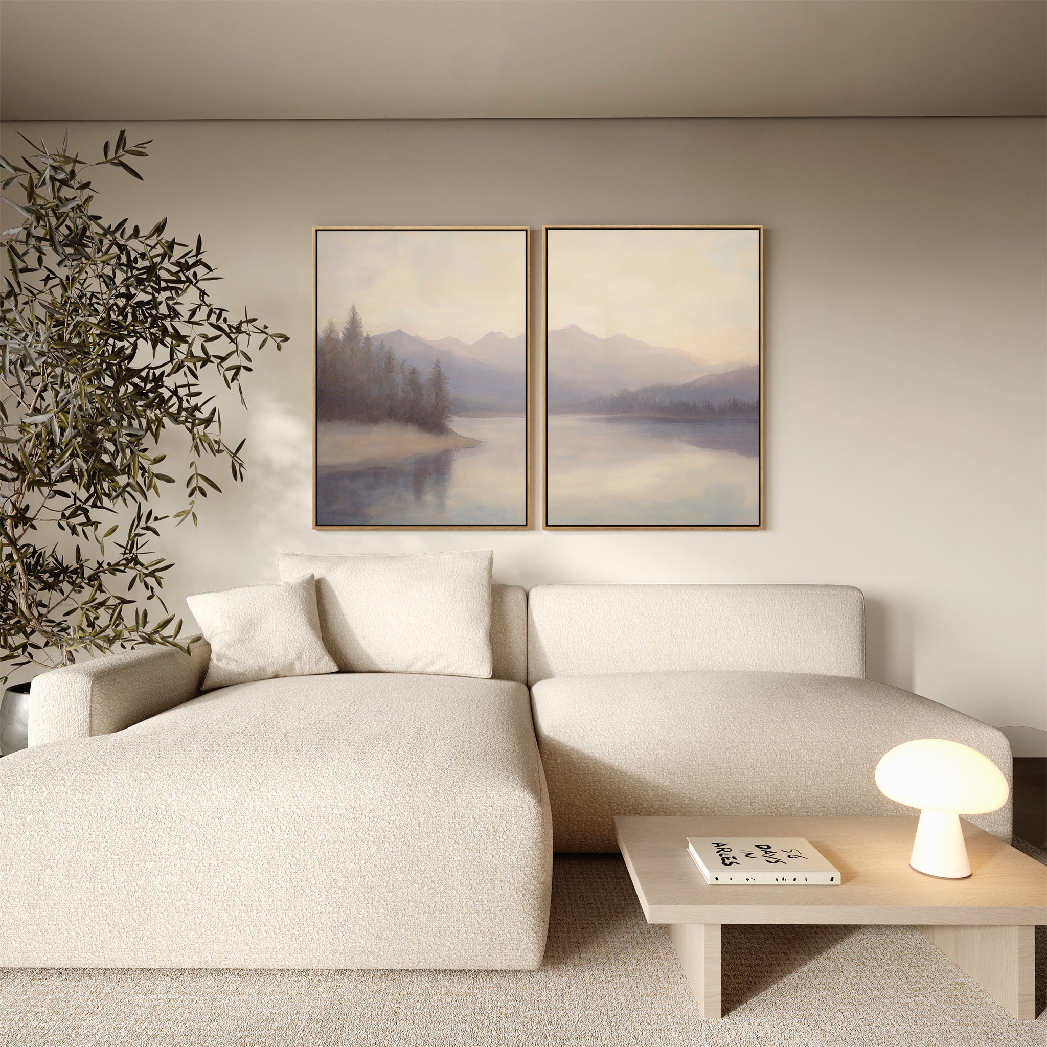 A minimalist living room features a light beige sectional, a small wooden coffee table with a lamp and book, a potted tree, and the Smoky Peaks Set of 2 landscape wall art canvases depicting mountains and water.