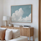 A modern living room displays a light wood sideboard with books, a lamp, and a bowl. Above it hangs the Soaring Peace Canvas Art, showing white clouds in a serene sky. Cream cushions from the sofa appear in the foreground.