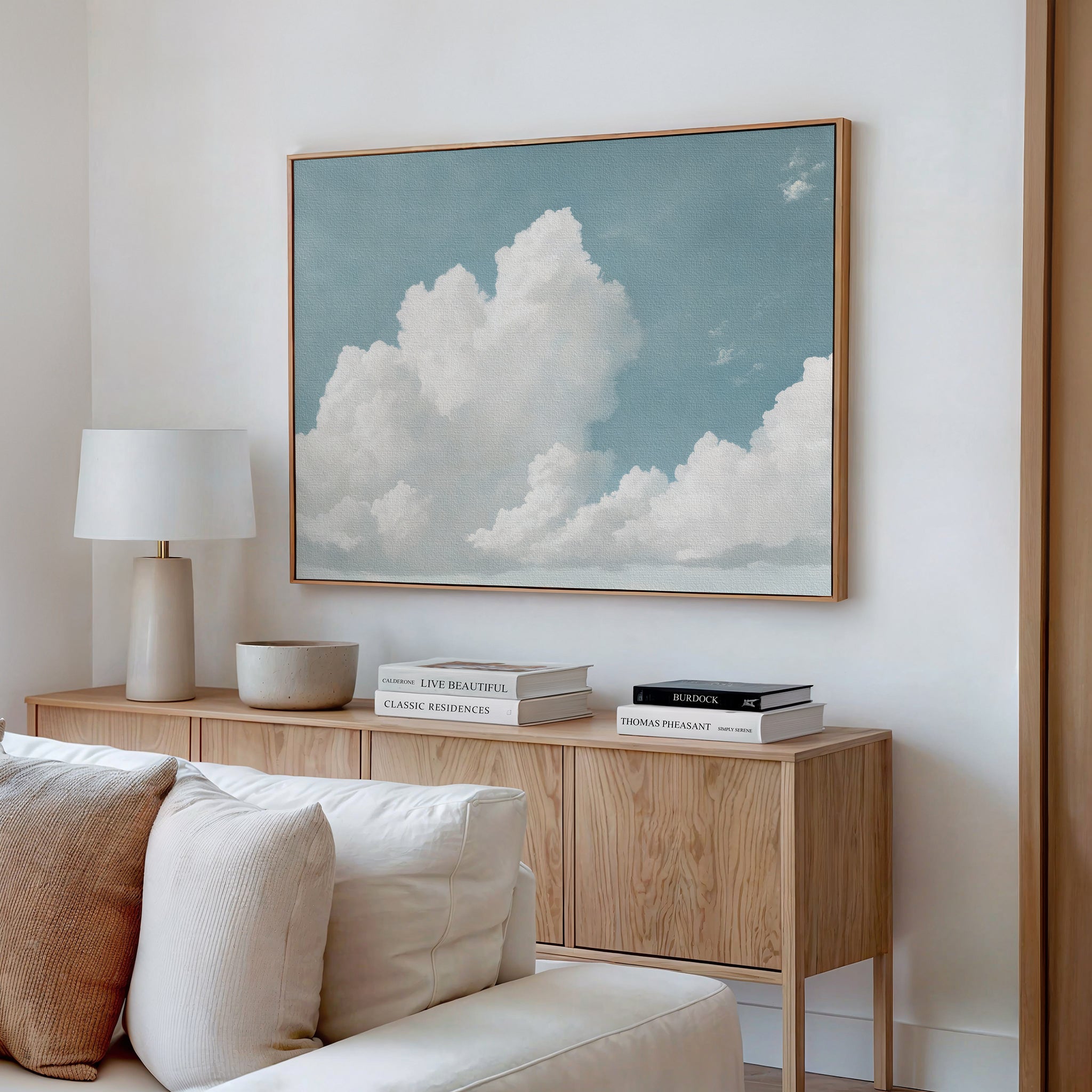 A modern living room displays a light wood sideboard with books, a lamp, and a bowl. Above it hangs the Soaring Peace Canvas Art, showing white clouds in a serene sky. Cream cushions from the sofa appear in the foreground.