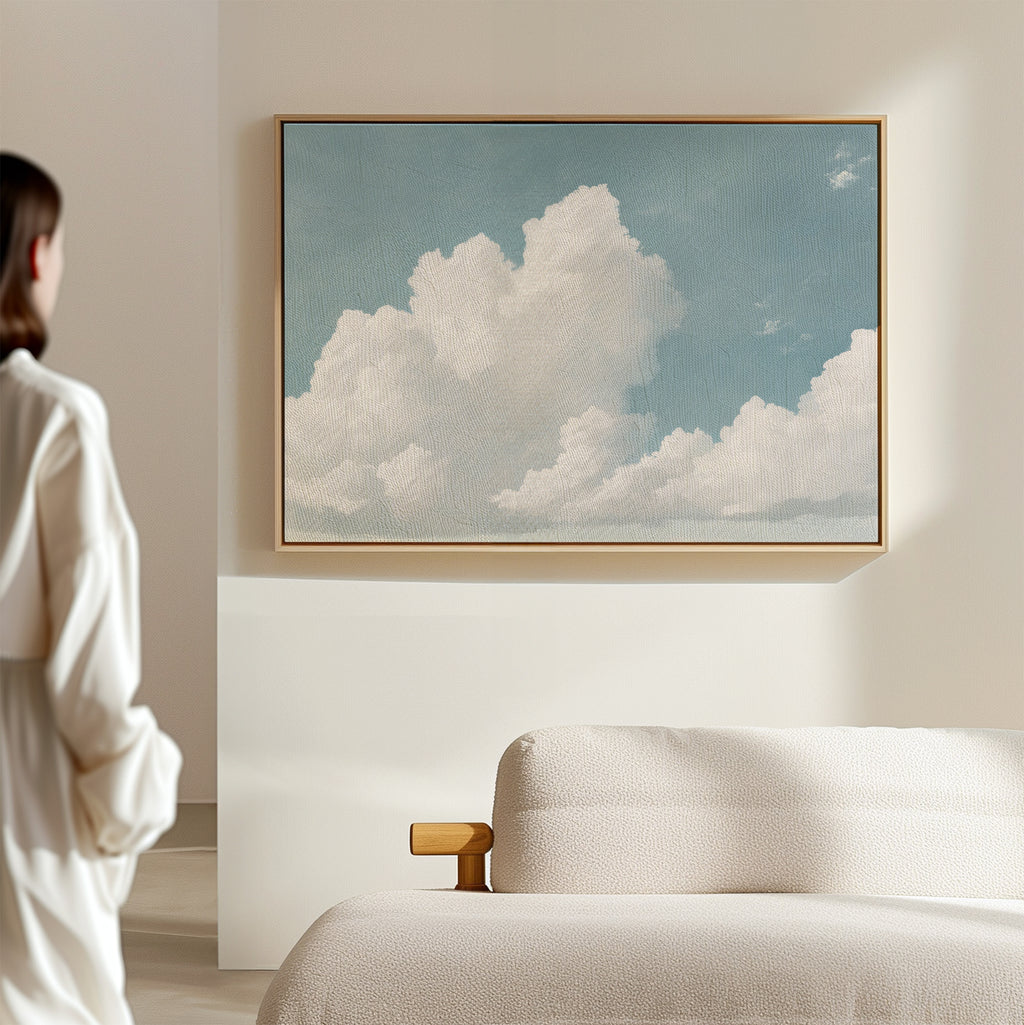 A woman stands in a minimalist room, gazing at the Soaring Peace Canvas Art featuring white clouds in a serene sky above a beige sofa. Soft natural light and vintage accents enhance the calm, tranquil atmosphere.