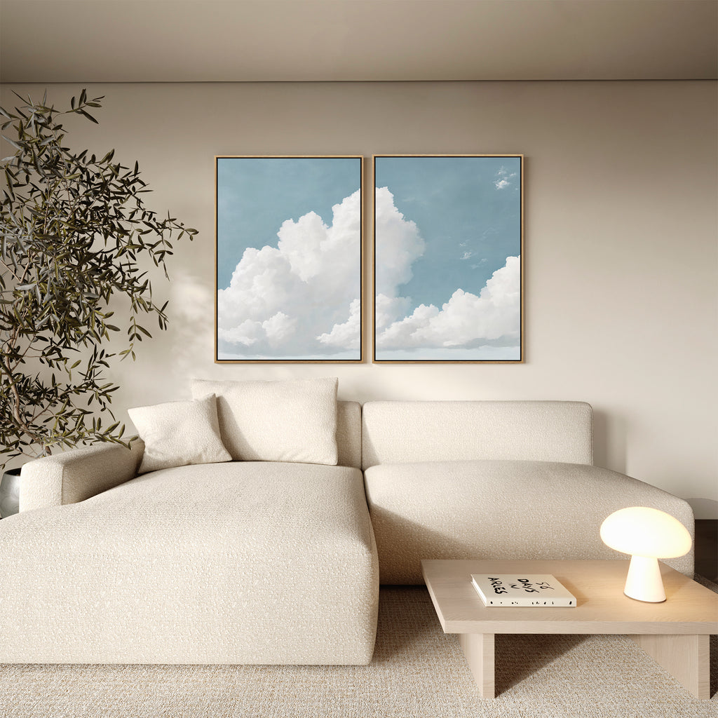 A modern living room with a beige sectional sofa, small wooden coffee table with books and a round lamp, vintage decor accents, a leafy plant, and the Soaring Peace Set of 2 cloud wall art prints on a light-colored wall.