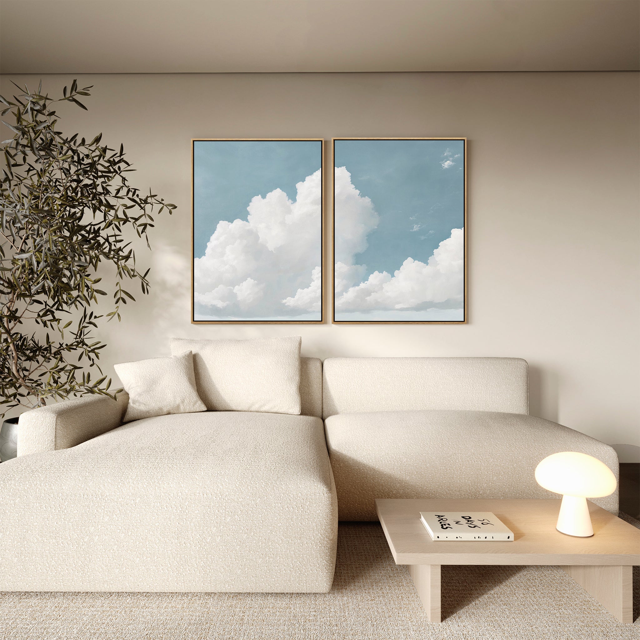 A modern living room with a beige sectional sofa, small wooden coffee table with books and a round lamp, vintage decor accents, a leafy plant, and the Soaring Peace Set of 2 cloud wall art prints on a light-colored wall.
