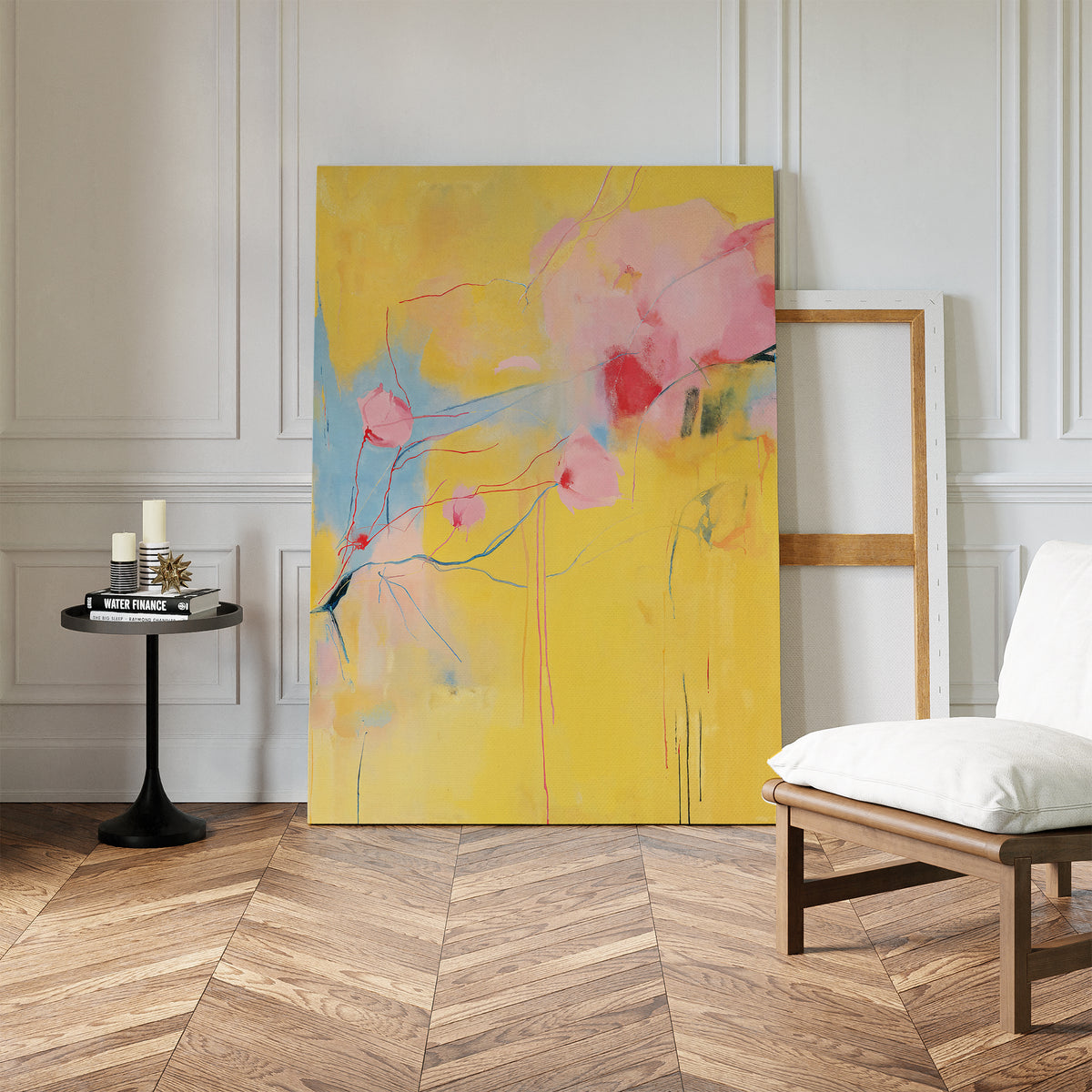 The Soft Bloom Abstract Canvas Art, with yellow, pink, and blue tones, leans against a white wall next to an empty frame. A black side table with candles adds charm beside a cozy white cushioned chair—perfect for uplifting your living room decor.