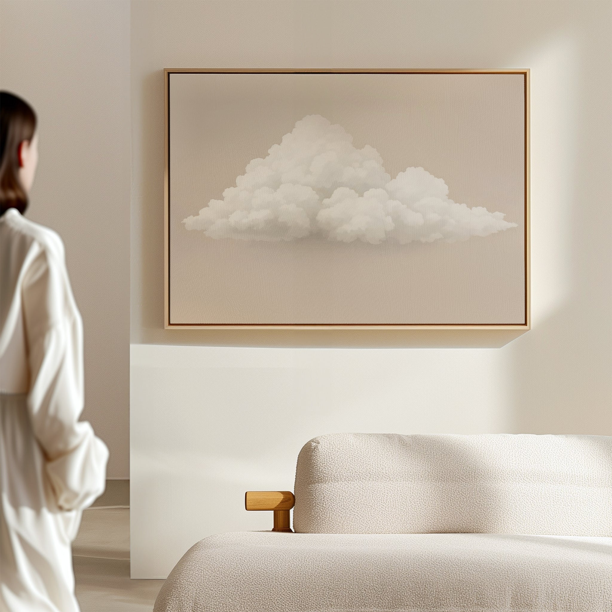 A person in white stands in a minimalist room, gazing at the Soft Drift Canvas Art—a fluffy white cloud on a beige wall above a light sofa—adding subtle charm and neutral tones to the space.