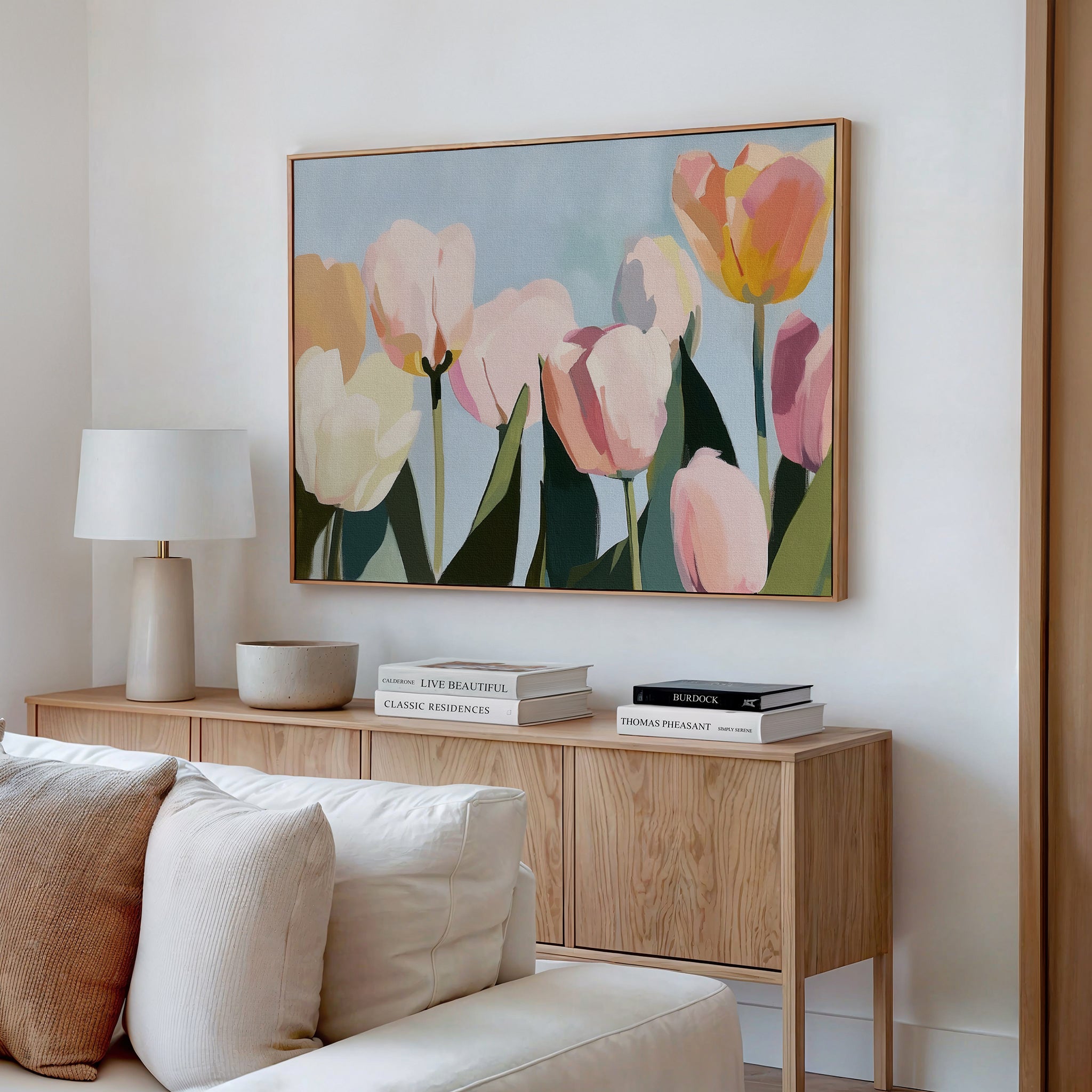 A bright, modern living room with a light wood sideboard, white lamp, books, and bowl features the Soft Field Canvas Art in pastel colors hanging above a cream sofa on a white wall.