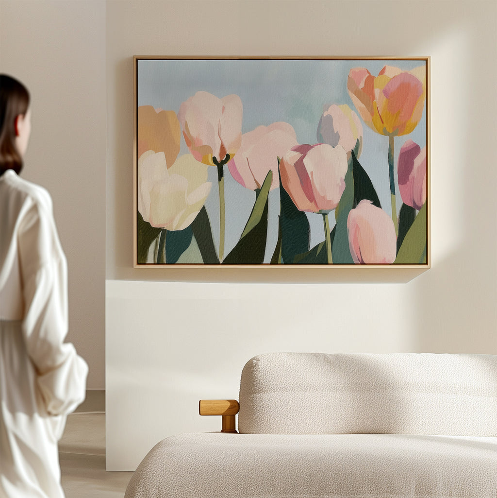 A woman in a white shirt stands in a softly lit room, admiring the Soft Field Canvas Art—a large framed painting of pastel tulips—hanging above a textured light sofa on a beige wall.