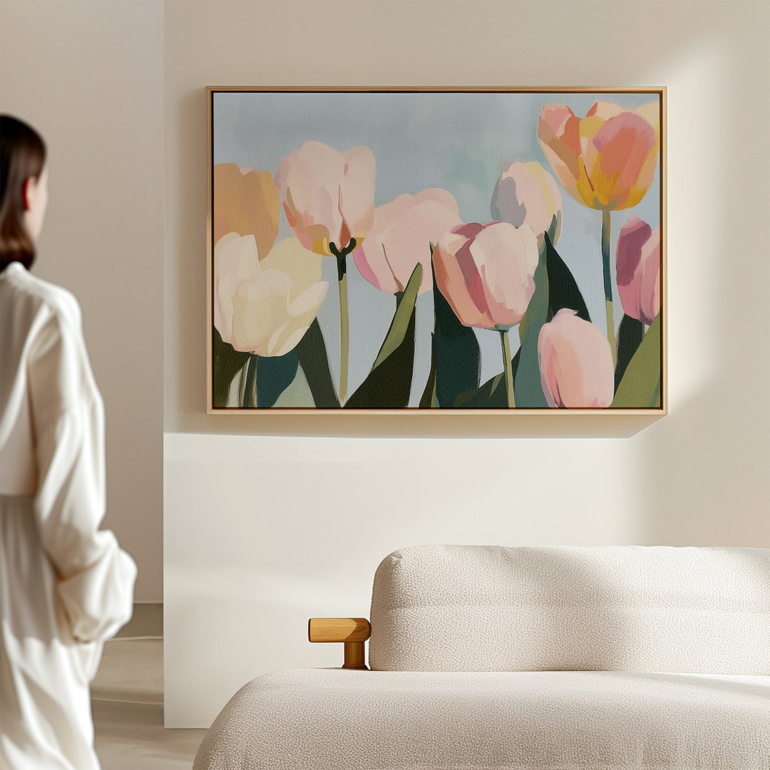 A woman in a white shirt stands in a softly lit room, admiring the Soft Field Canvas Art—a large framed painting of pastel tulips—hanging above a textured light sofa on a beige wall.
