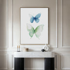 The Soft Flight Canvas Art, a minimalist piece with blue and green watercolor butterflies, is framed and displayed above a marble-topped Nordic Park console decorated with candles, books, and a sculptural white vase.