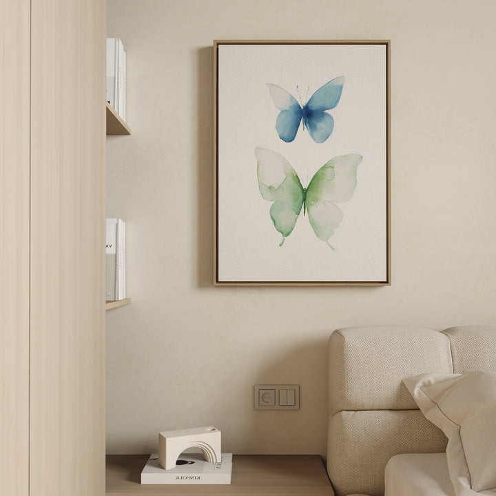 A minimalist living room wall features the Soft Flight Canvas Art—Nordic-inspired watercolor butterflies, with a blue butterfly above a green one—framed and displayed beside beige shelves of books and a cream sofa.